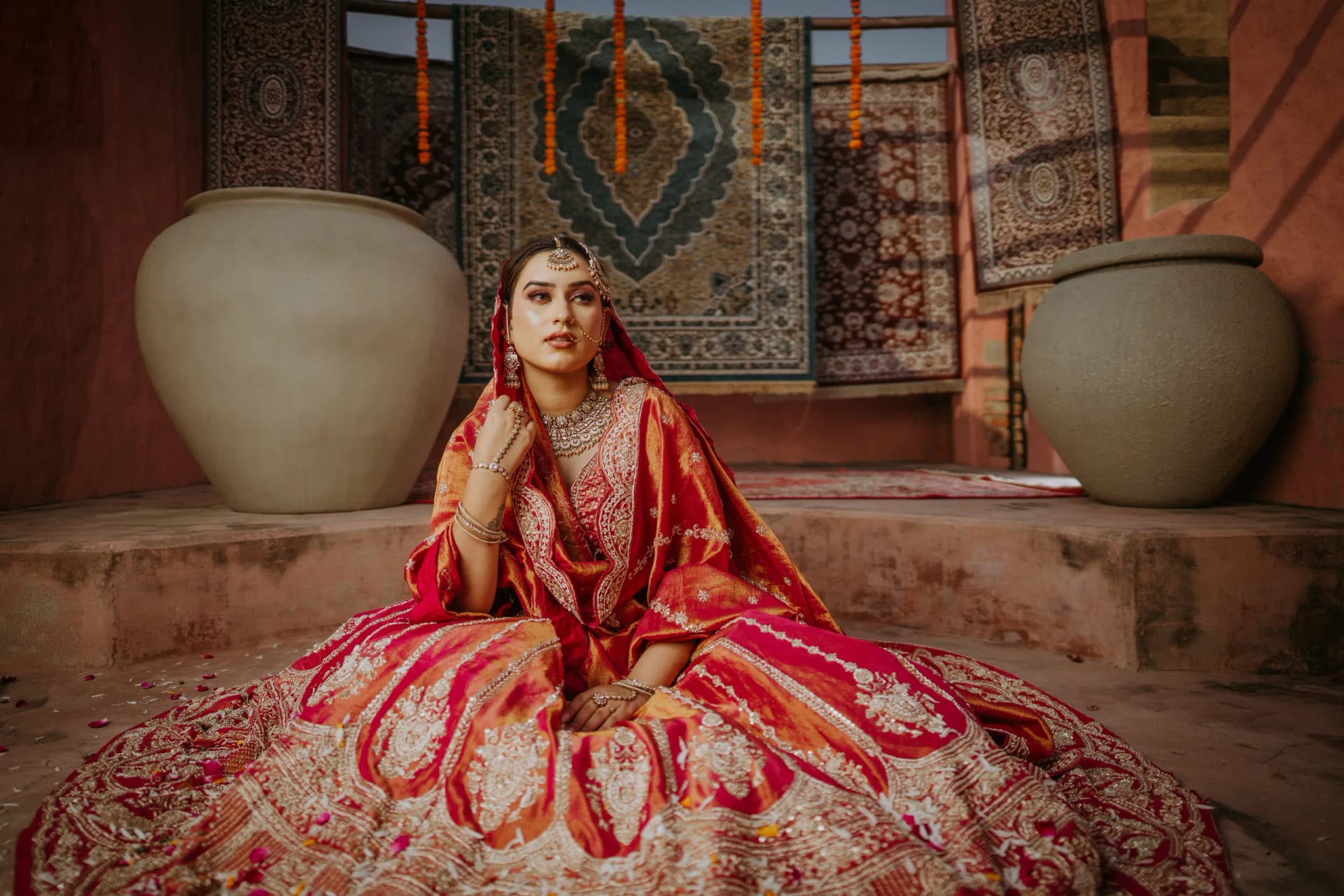 A bride in a rose-red and gold hand-embroidered lehenga, seated in a terracotta courtyard hung with woven rugs and marigold garlands
