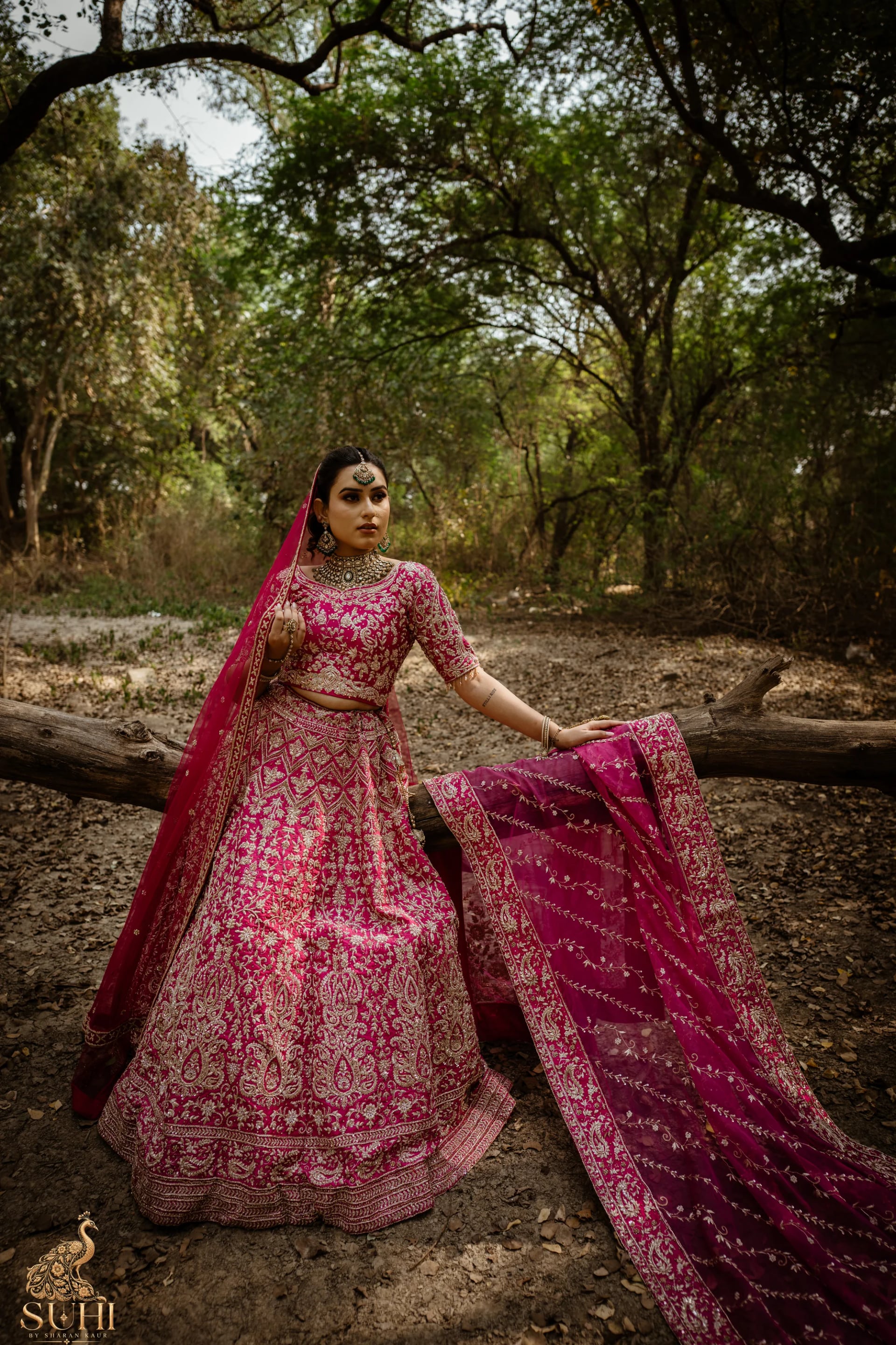 Bride in the RAANI GUL rose-red bridal lehenga standing in a forest clearing, the dupatta spread beside her